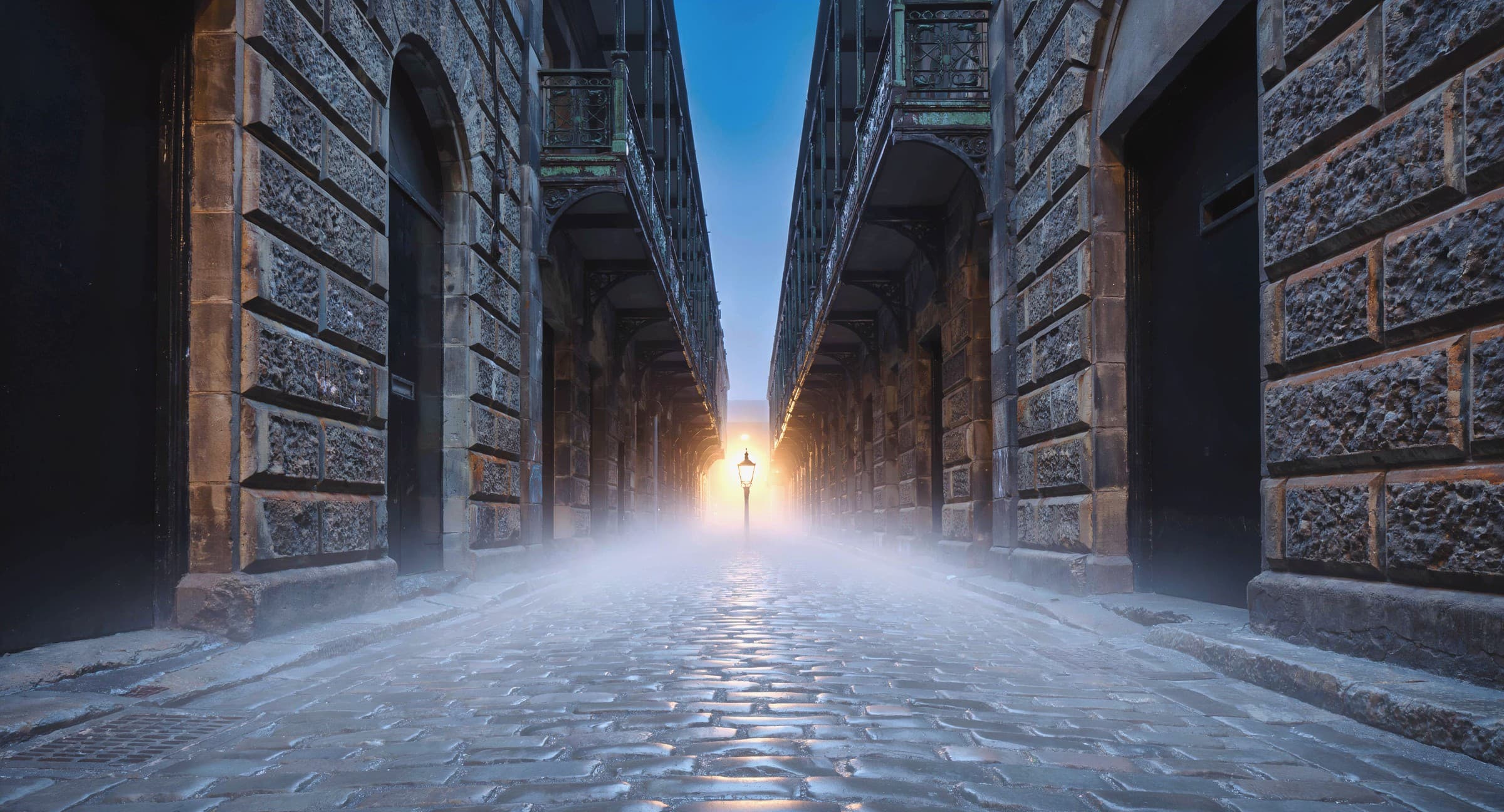 A wet bluestone Melbourne laneway at dawn, Victorian-era warehouse facades on either side, a single brass lamp at the far vanishing point glowing through low fog.