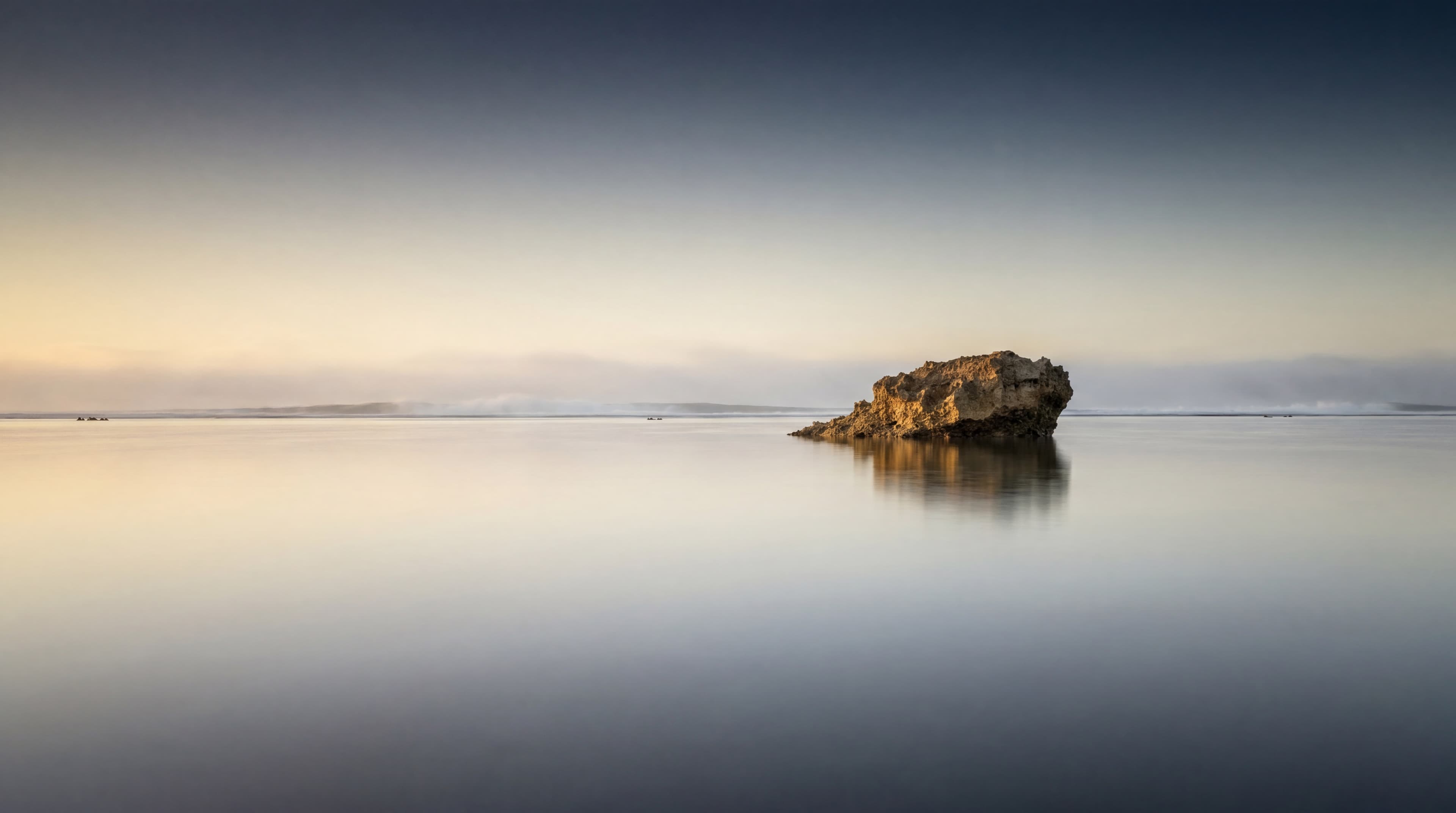 A Pacific atoll at first light, a single stone outcrop emerging from a mirror-still lagoon under a brass-to-navy dawn sky.