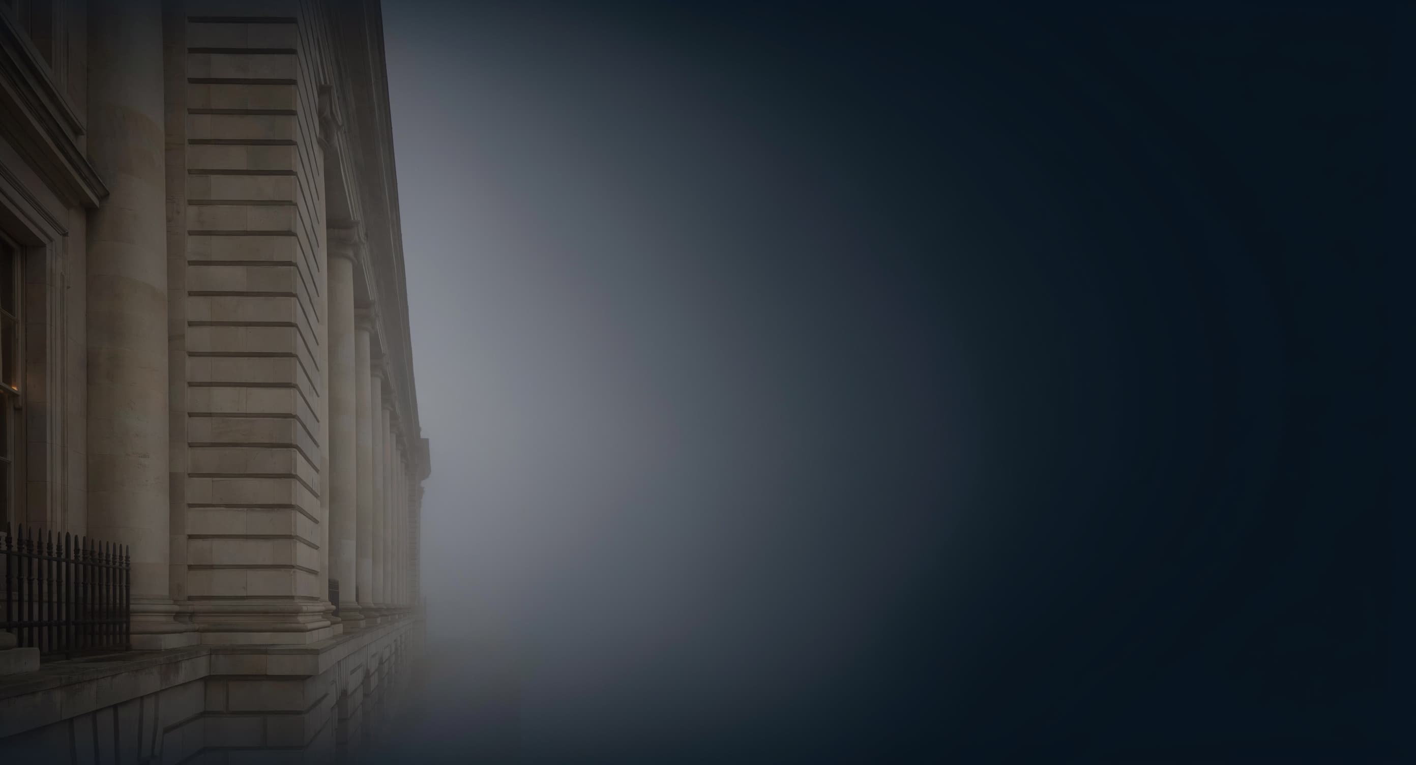 A Georgian institutional stone facade in heavy fog at dusk, asymmetric, a single warm sash window glowing at the frame's edge.