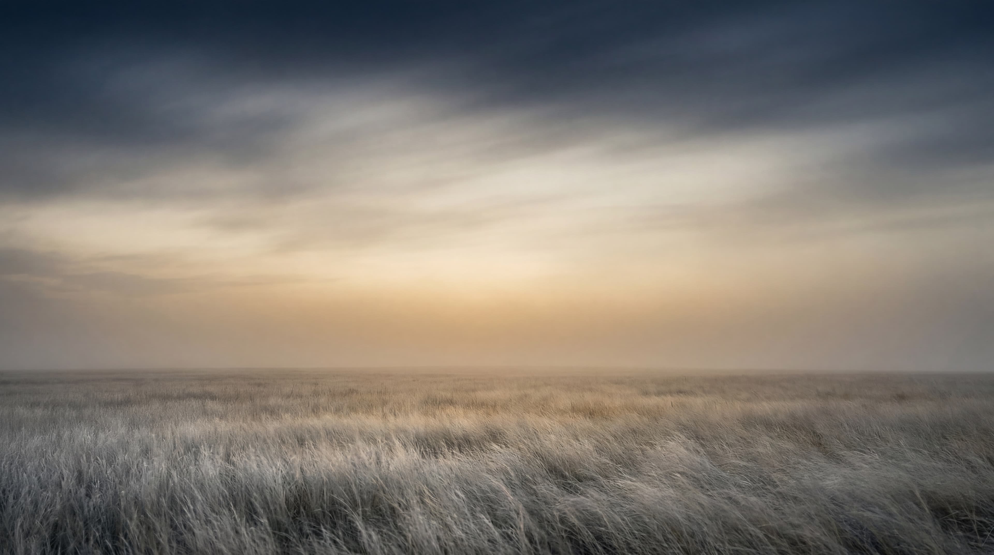 A long-exposure high-plains horizon at first light, brass-warm glow bleeding into deep navy.